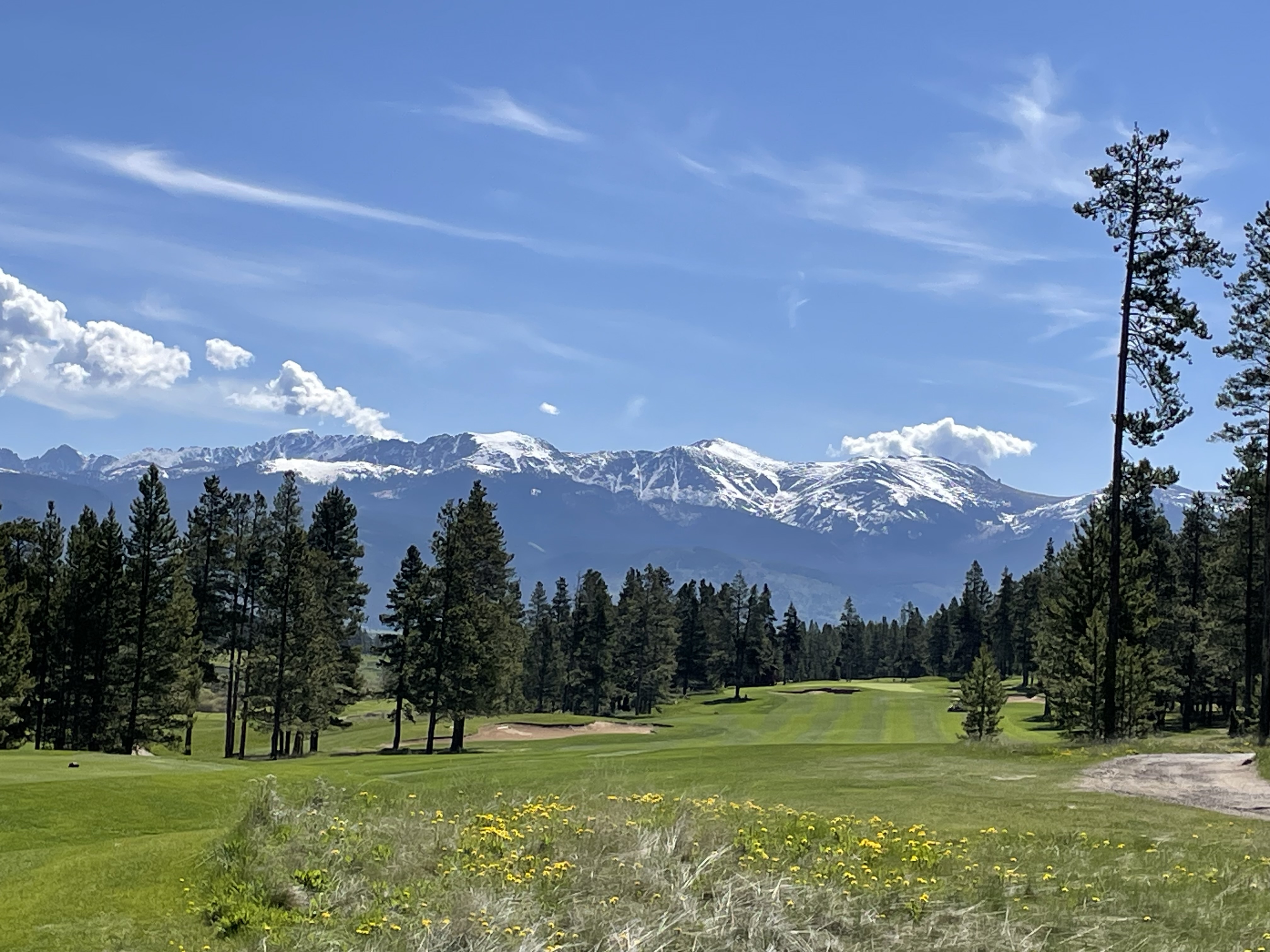 A golf fairway surrounded by pine trees beneath snow-capped mountains and a blue sky
